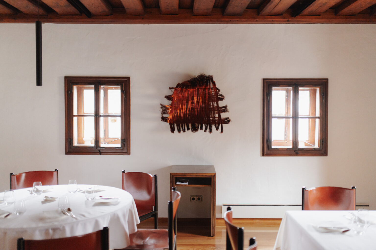 Gedeckte Tische im Mesnerhaus Gourmet-Restaurant zwischen zwei Fenstern und einem roten Wandkunstwerk
