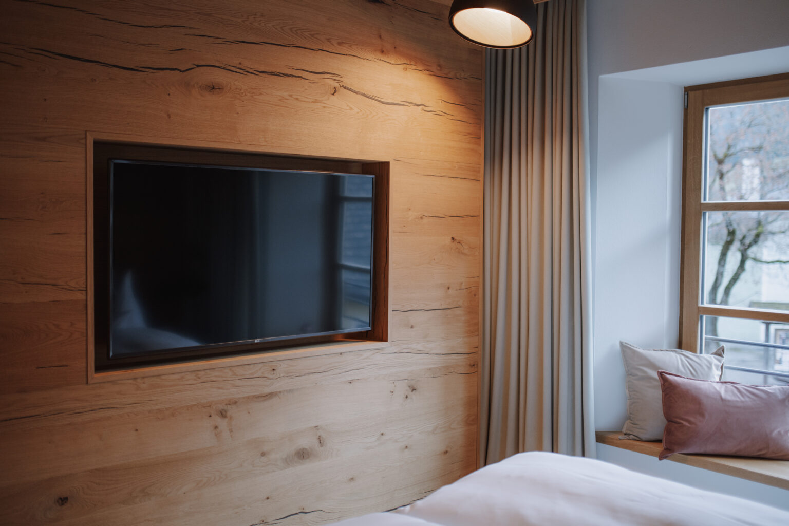Fernseher an einer Holzwand und Fensternische in einem Zimmer des Mesnerhaus Boutique-Hotels